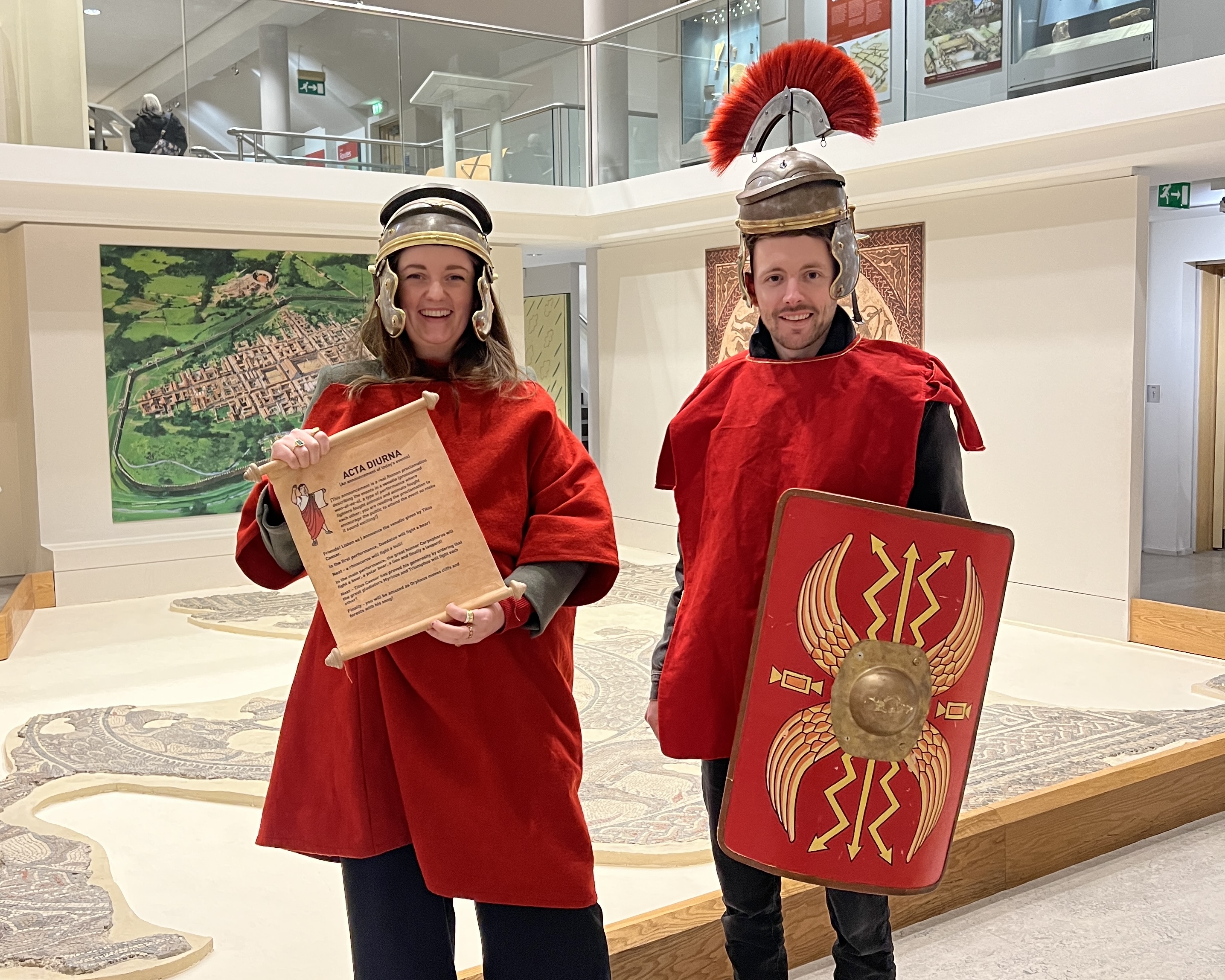 A man and woman dressed in red roman costumes pose beside an roman floor mosaic at the Corinium Museum in Circencester
