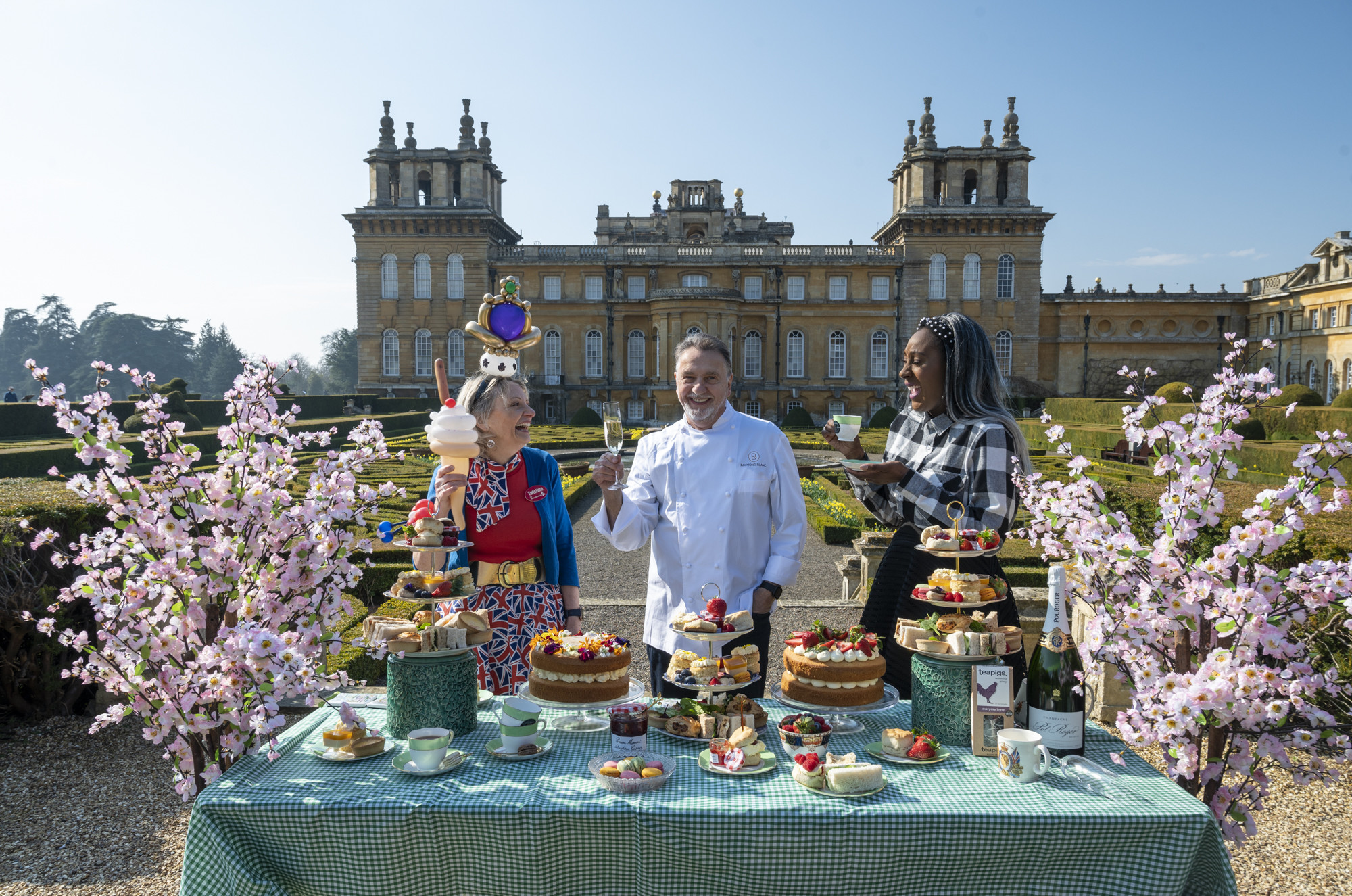 Chef and two guests stood at a display table in front of Blenheim Palace, with food and cakes on the table in front of them