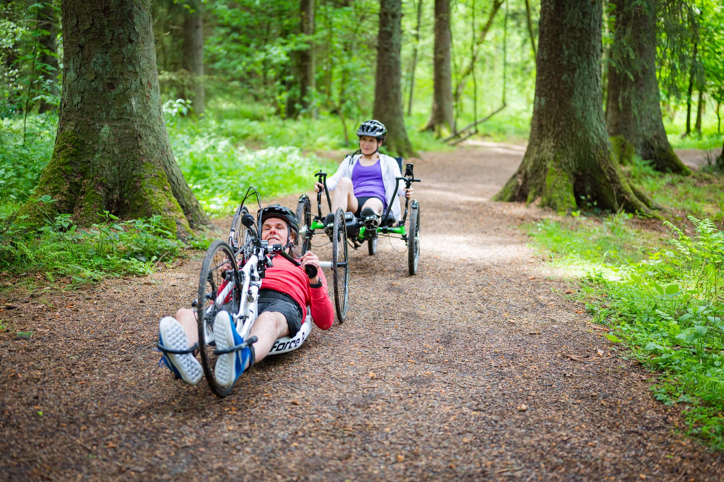 Woman on recumbent trike and man on recumbent hand crank cycle ride through forest