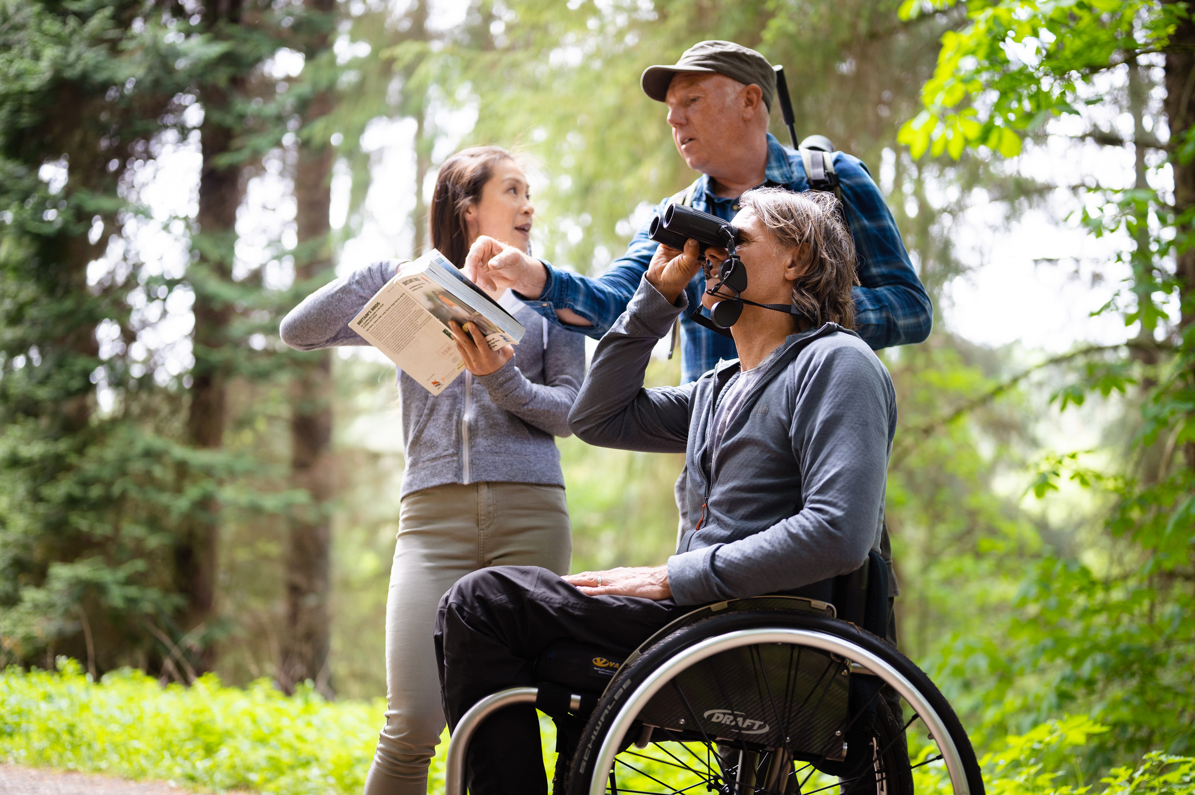 Man in checked shirt with telescope is telling two people, a woman and a man using binoculars and a wheelchair about the forest bird song