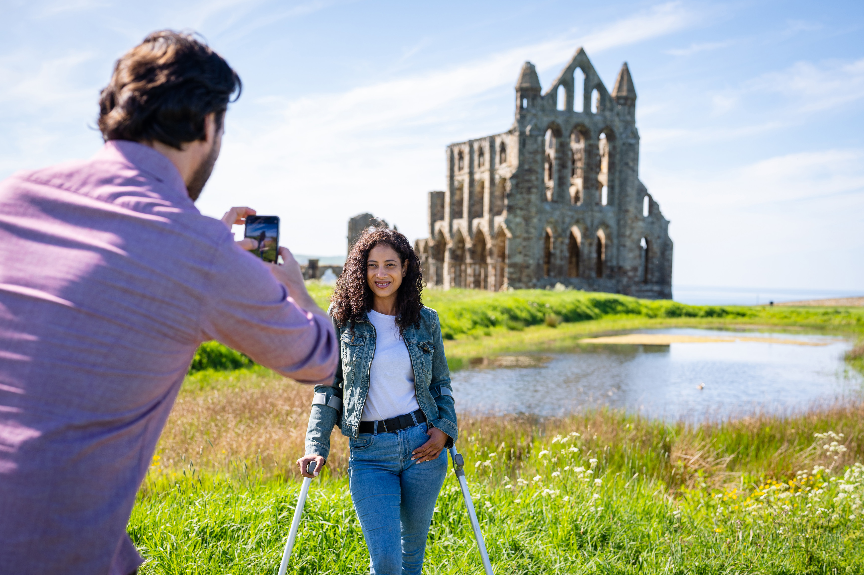 Man taking a photograph of woman, using crutches, with ruins of Whitby Abbey and lake in the background