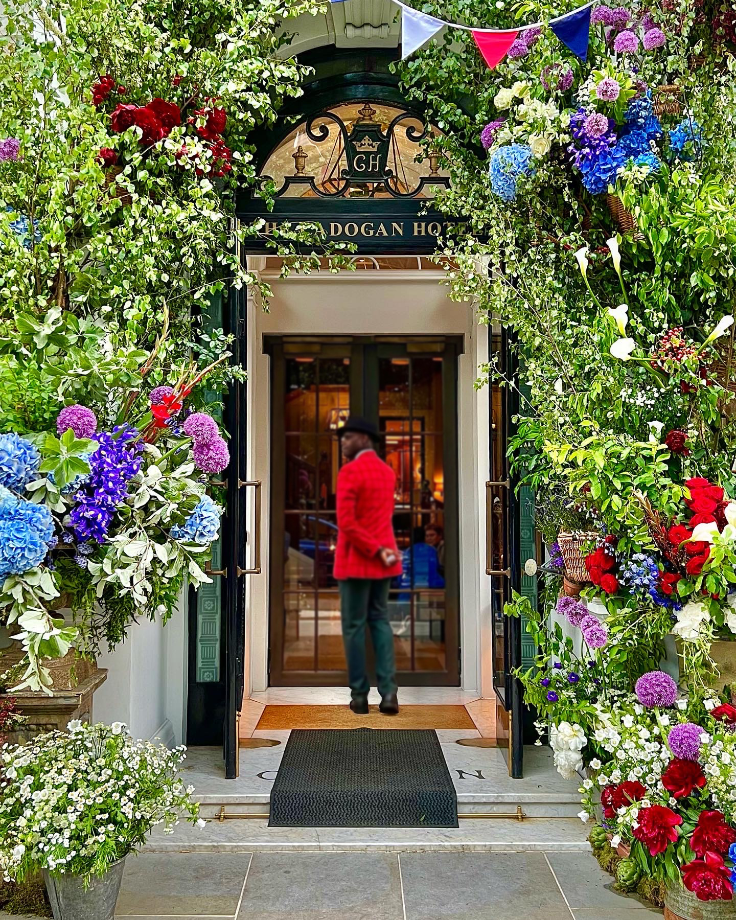 Colourful flowers surrounding the door to The Cadogan Hotel with a doorman in a red jacket about to enter 