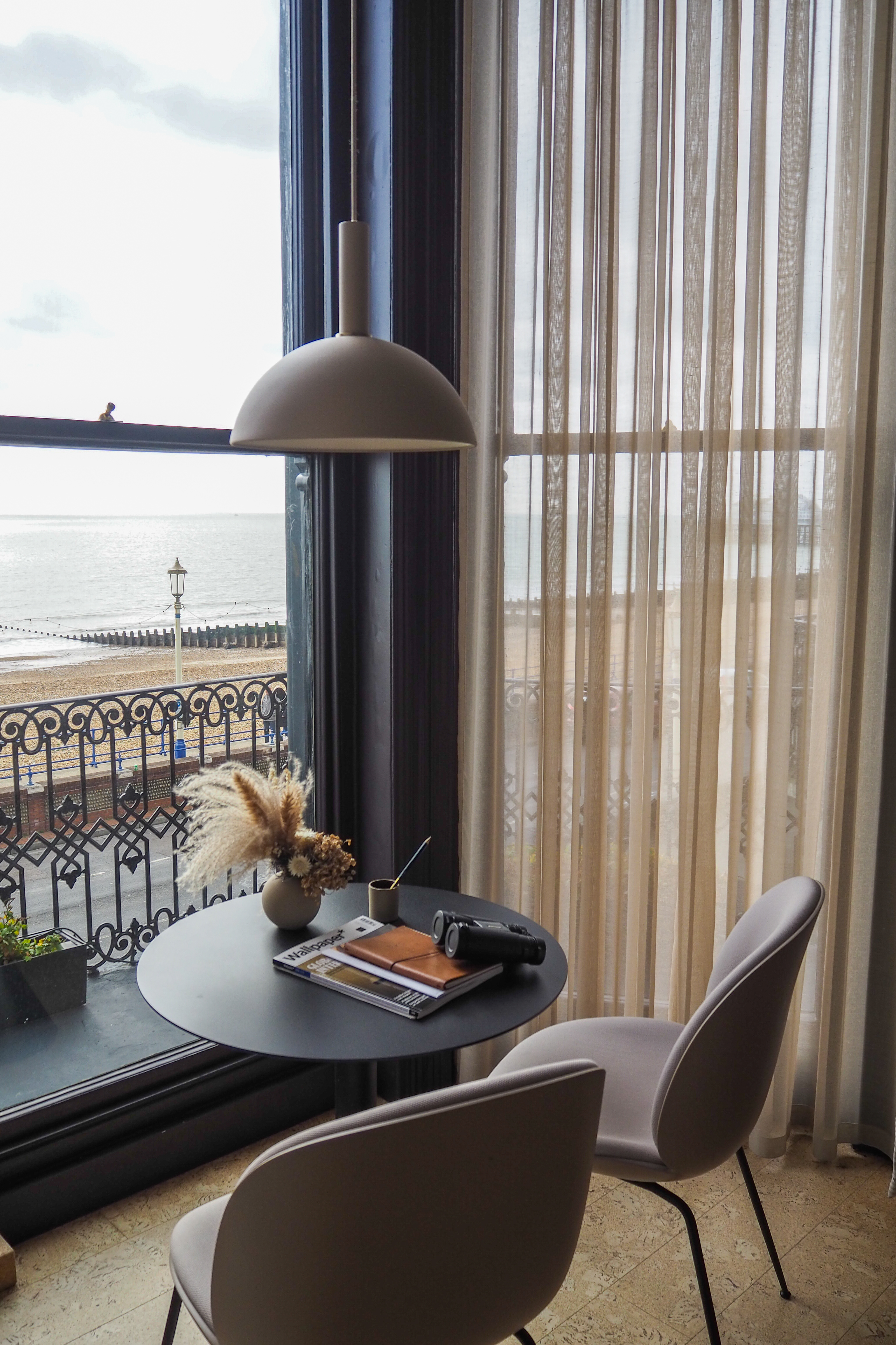 A hotel room with a table and chairs infant of a large bay windows revealing Eastbourne Beach