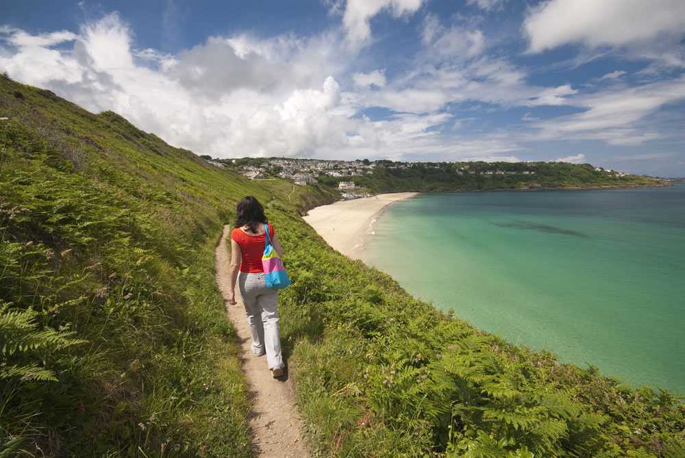 A woman strolls along a grass-flanked coastal path next to teal-coloured water in Cornwall