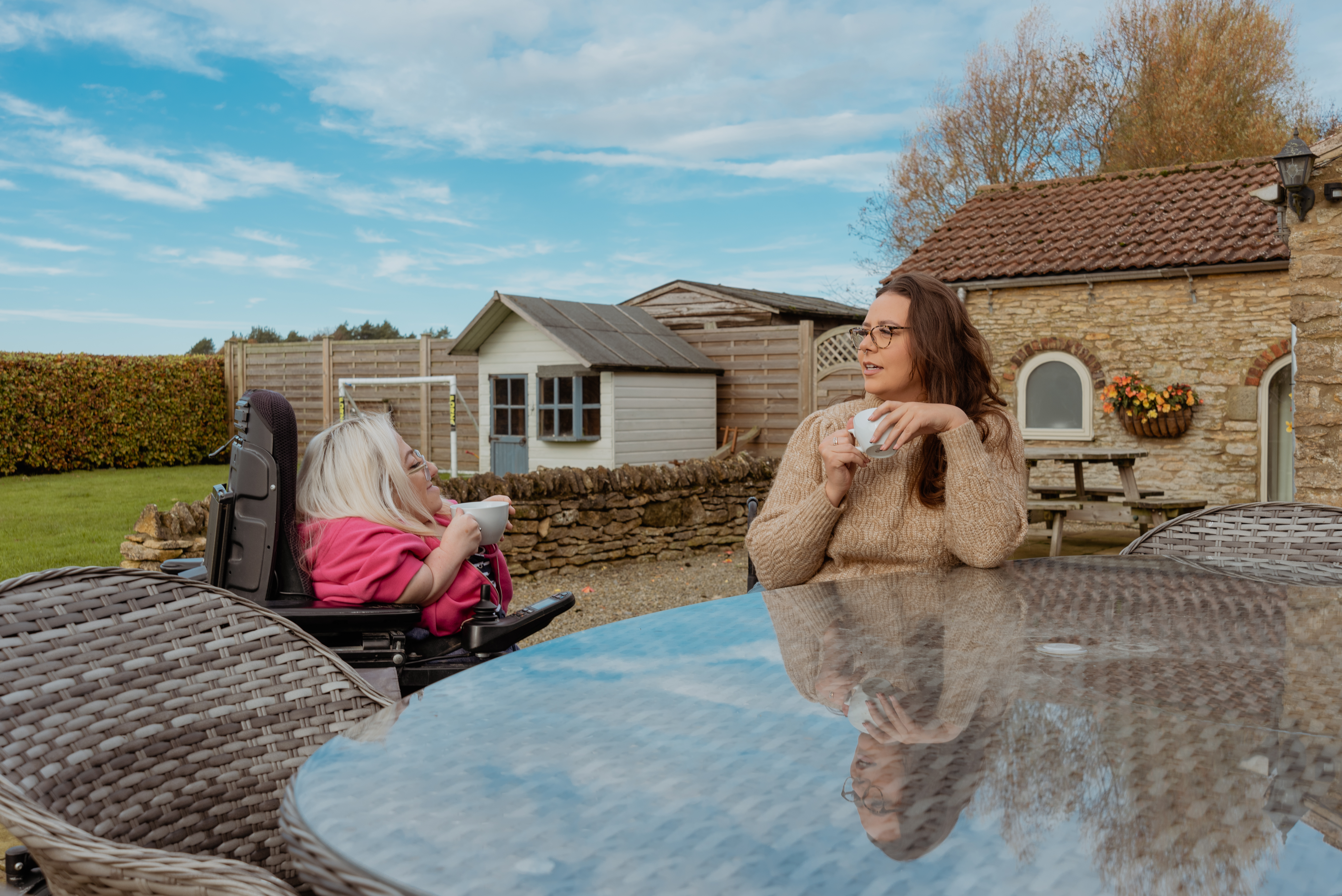 Two women in wheelschairs sit in a garden enjoying a hot drink as a bright blue sky reflects in glass table top that are sat at