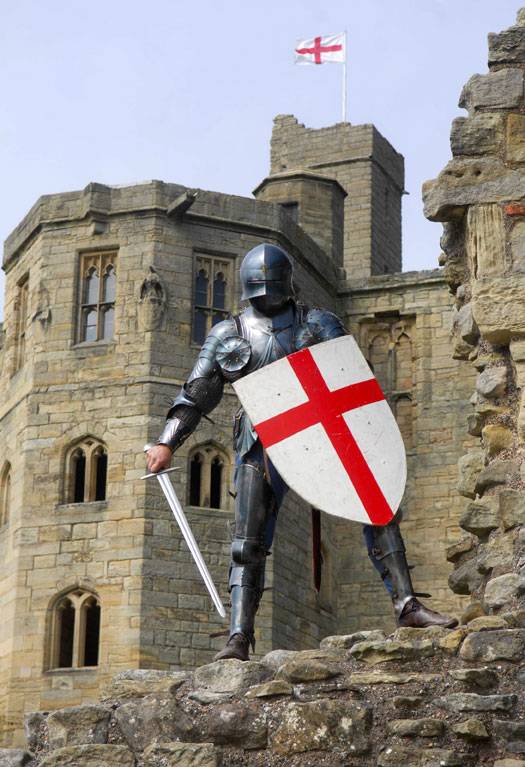 A knight in armour holding a St George's Cross shield stands in front of Warkworth Castle