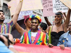 Parade goers during Pride in London in July 2019. A black guy wearing a colourful blazer and with a colourful headpiece carries the LGBTQ+ flag wit a black woman, who wears a denim jacket on the top of a striped t-shirt