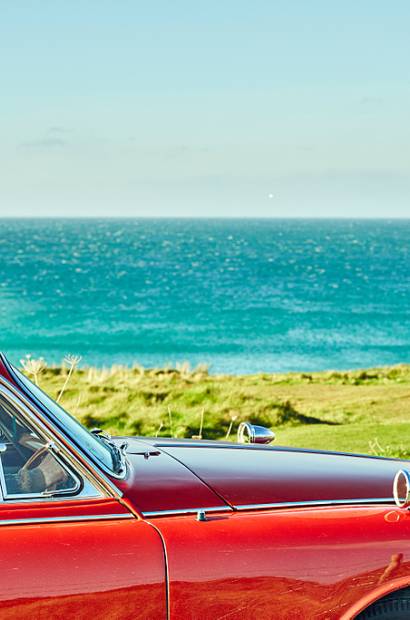 A couple on the North Cornish coast, walking along the clifftops. A red Classic Jaguar car parked.