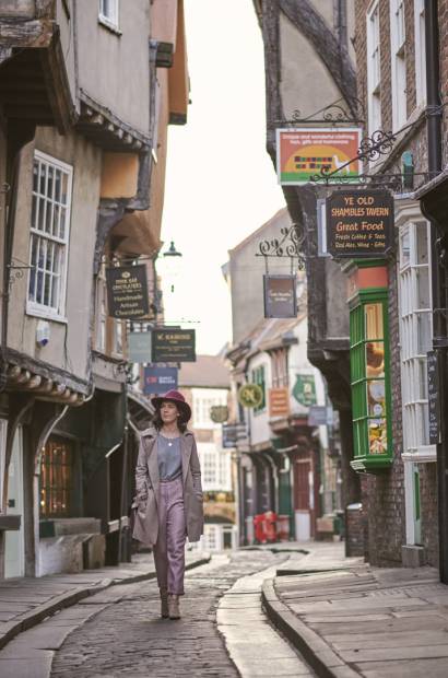 Woman wearing trench coat and pink hat walking through narrow historic street of York, North Yorkshire, England.