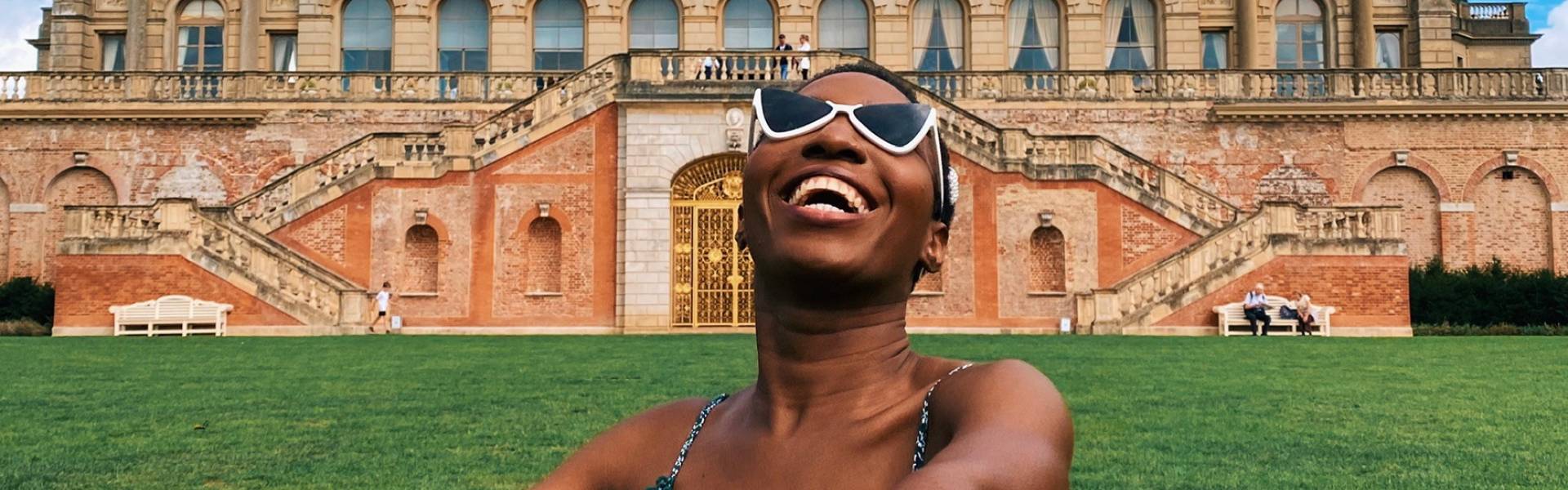 Smiling woman, wearing sunglasses and sundress, outside Cliveden House - a large stately home.