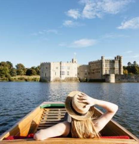 A woman in a summer hat sits in a wooden boat with Leeds Castle in the near distance