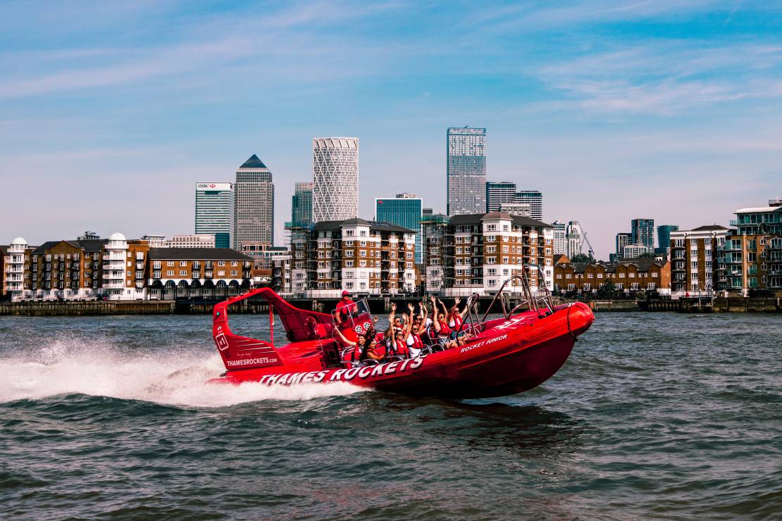 Passengers cheering on a red speed boat on river Thames in front of sky scrapers at Canary Wharf