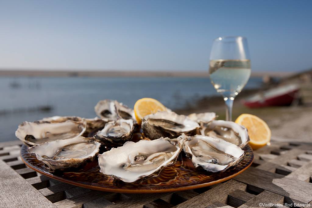 A platter of open shucked oysters with garnish of fresh lemons. A glass of chilled white wine. View out over the beach and sea.