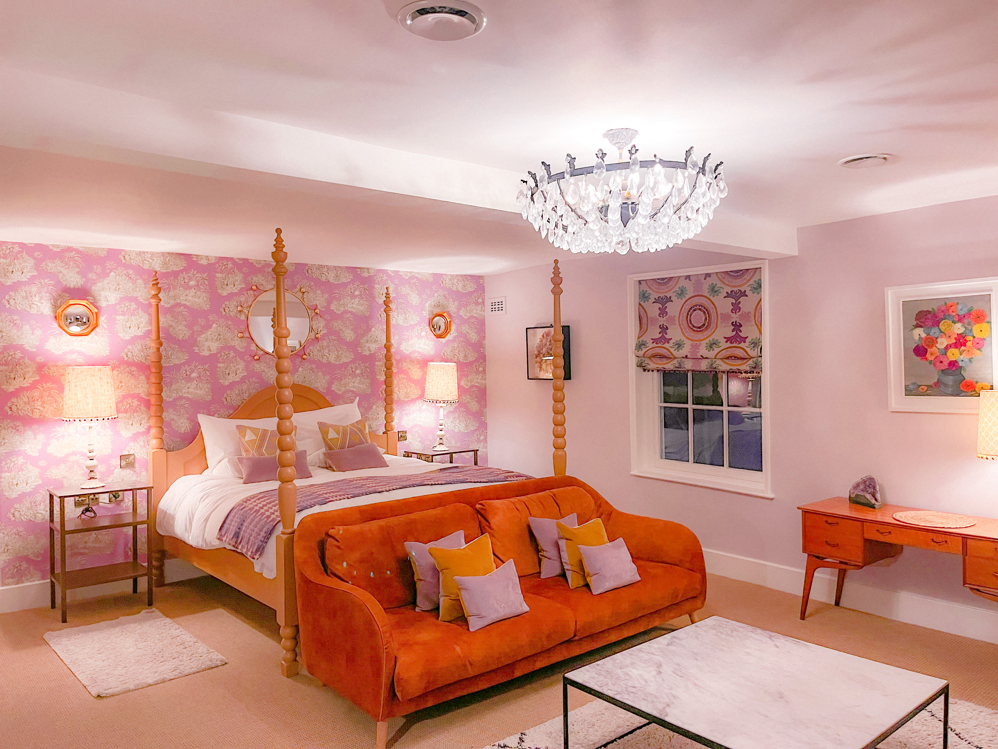 A luxury hotel room with colourful wall paper, a modern four poster bed and a bright orange sofa perched at the end of it