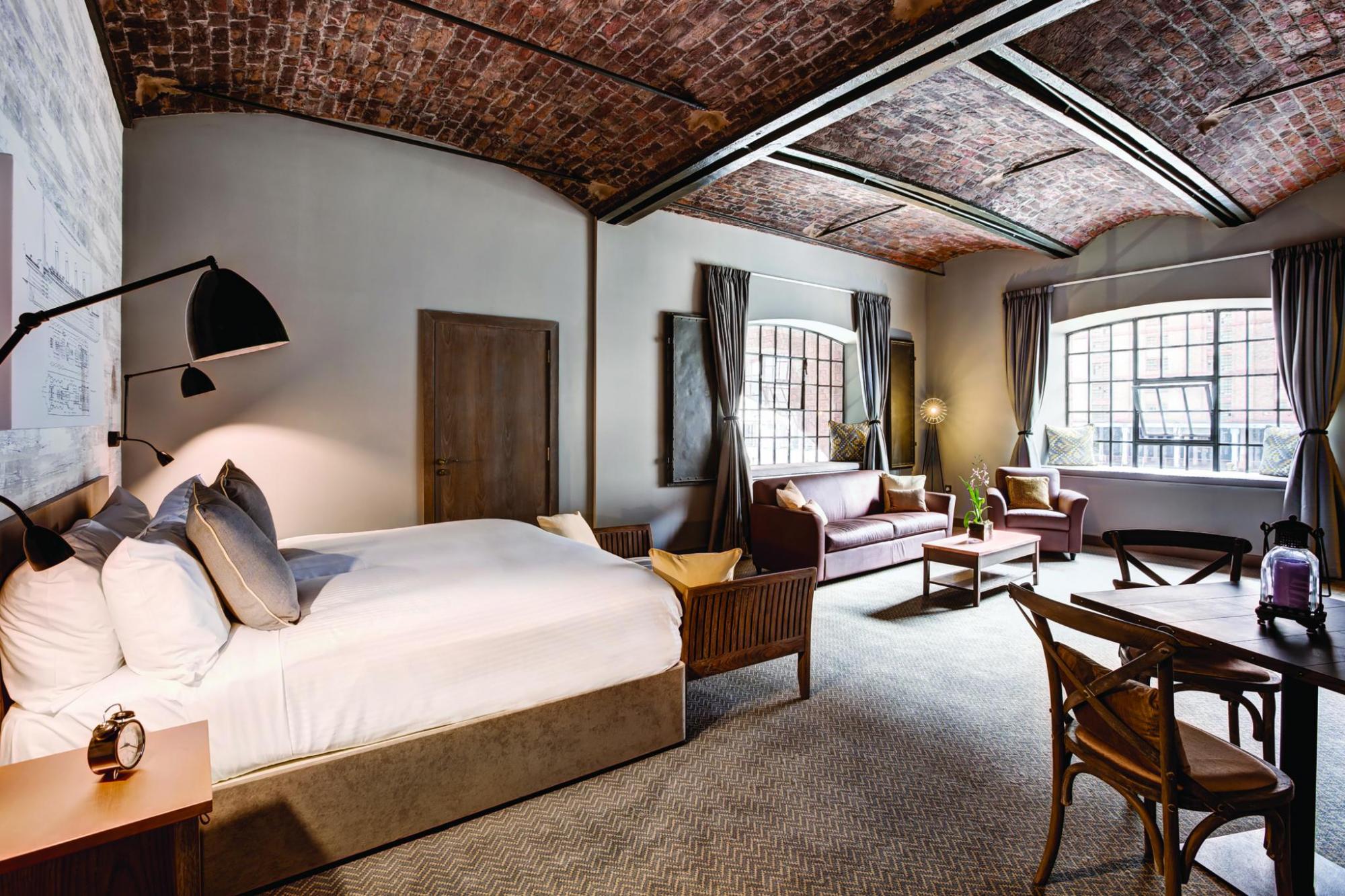 Large hotel room with exposed brick ceilings and steel beams, with big window letting in light