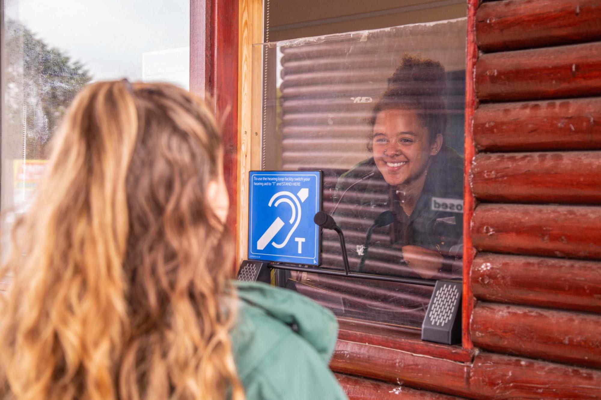 Young girl at ticket hut, speaking with smiling staff member behind window with blue hearing sticker visible