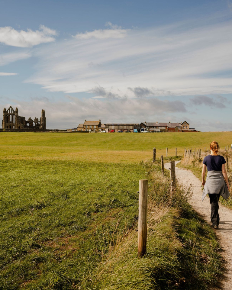 Woman walking along a coastal path towards a ruined abbey