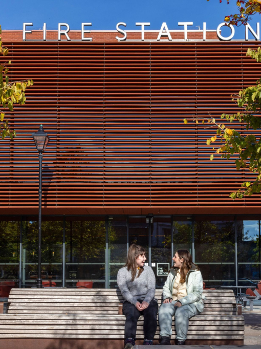 Two people sat on a bench outside of a Fire Station auditorium, framed by leafy trees.