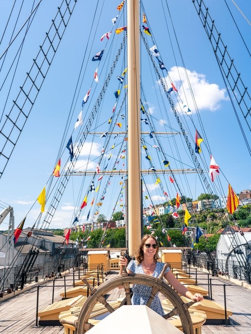 Smiling young woman in sunglasses at the wheel of a ship docked on land with flags on guide ropes against blue sky