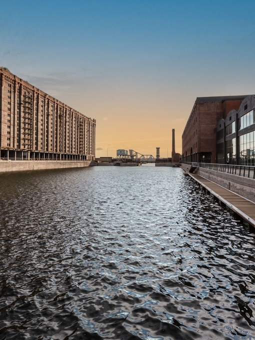 A view of a dock looking across the water. To the left stands a traditional brick built dockside building, to the right is a modern building made from glass and wood. 