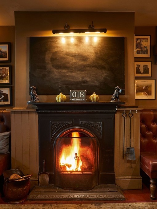 A fireplace with a lit fire flanked by two leather sofas with tables. The wall is covered in numerous framed artworks. The Duncombe Arms - Bronze award winner for the Pub of the Year at the VisitEngland Awards for Excellence 2023.