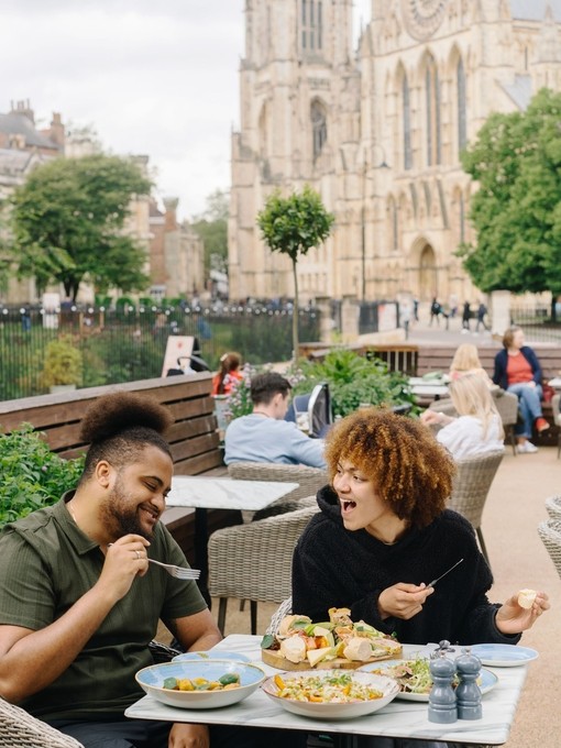 A man and a woman eat lunch outside a cafe with a cathedral in the background