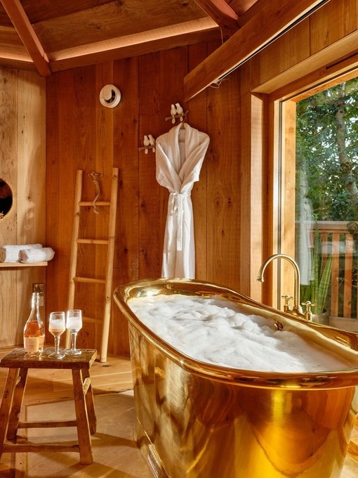 The interior of a wooden lodge, with a gold roll-top bath by the window at Sleepy Owl Devon.
