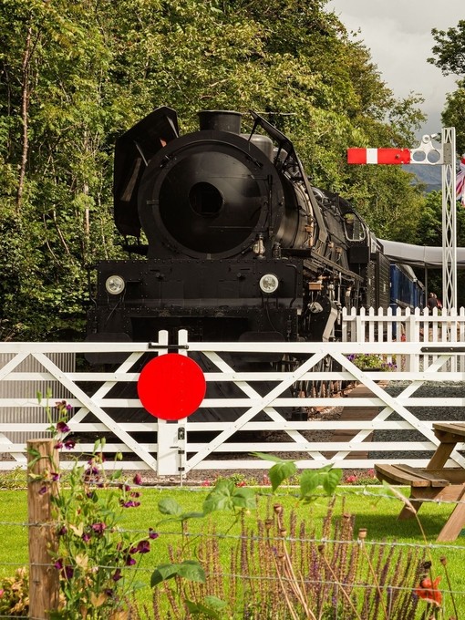 A front view of a parked steam train behind closed white level crossing gates, with a stone train station house to the right. In front is a green lawn with a picnic table. Bassenthwaite Lake Station - Silver award winner for New Tourism Business of the Year at the VisitEngland Awards for Excellence 2023.