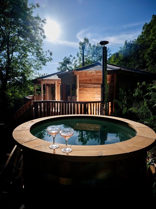 A wooden edged hot tub in front of the lit wooden lodge at Sleepy Owl Devon.