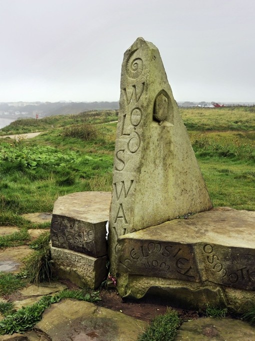 A marker stone with engravings stuck in the ground at the top of a moor with the sea beyond