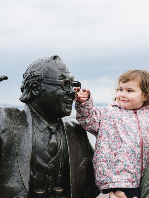 A young child and man stand next to a statue on the seafront, as the child reaches up to touch the statue’s face