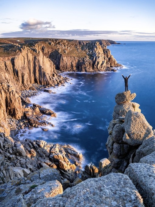 A man stands on a rocky peak looking over the rocky Cornish headland