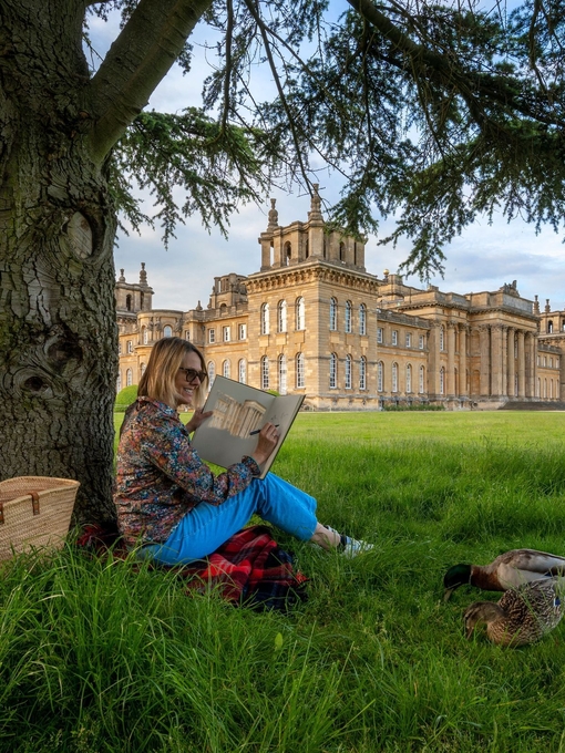 Woman sat under a tree sketching in the grounds of a large country estate