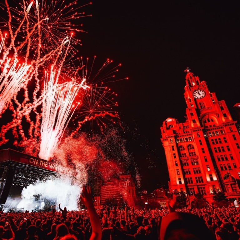 Crowds of people watching a band at a music festival as fireworks explode, in front of the Liver Building.