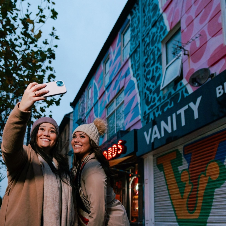Two women pose for a selfie in front of a row of shops