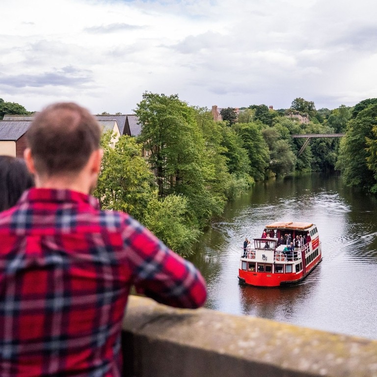 Two people stood on a bridge overlooking a tour boat sailing down a river in Durham