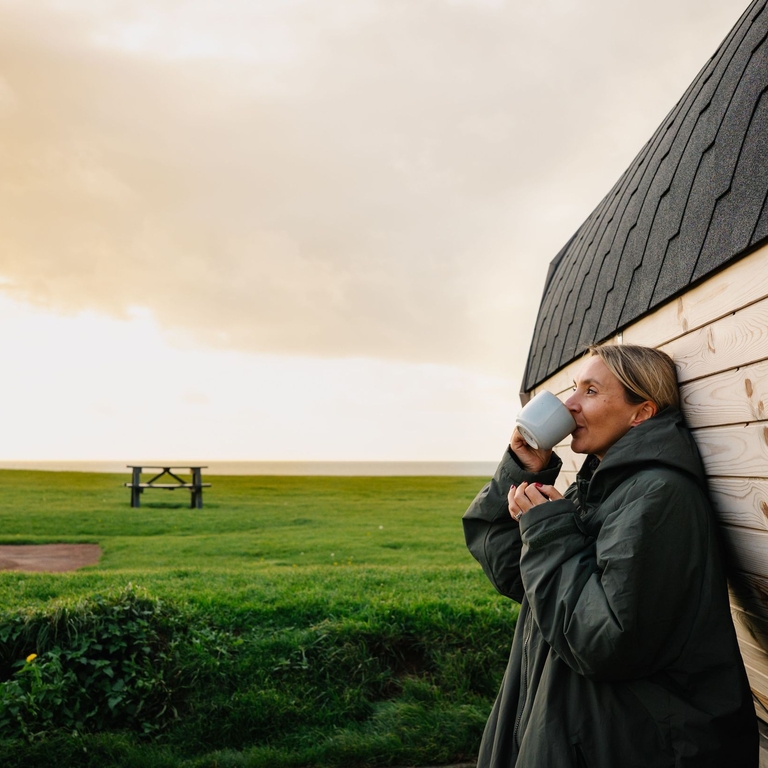 A woman drinks a hot drink while leaning against a sauna by the sea
