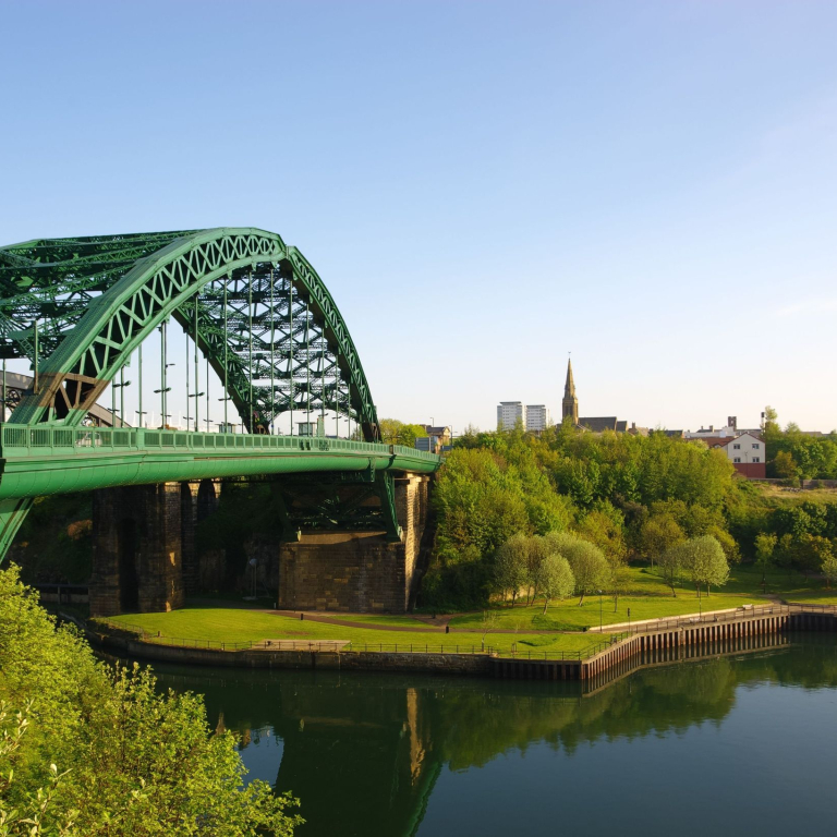 View of a green iron bridge crossing over a river, surrounded by trees and buildings