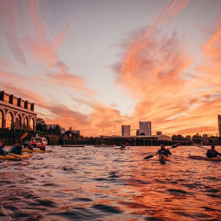 People kayaking in Thames against a red sunset sky and the buildings of London in the background