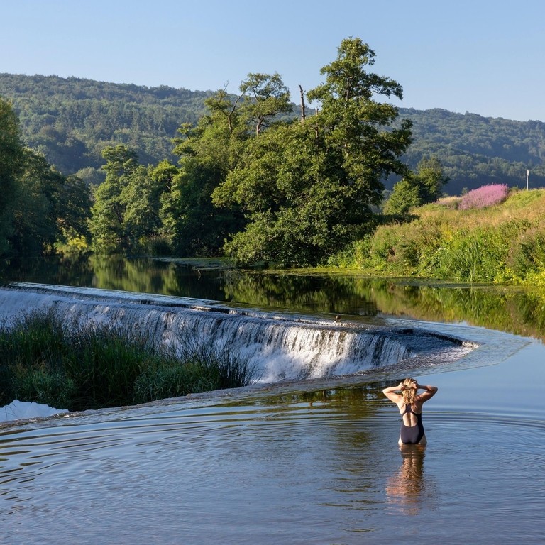 A female swimmer standing in the shallows preparing to swim in the a river above a weir with wooded hills in the distance