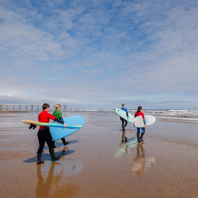 Four people walk toward the sea wearing wetsuits and carrying surf boards
