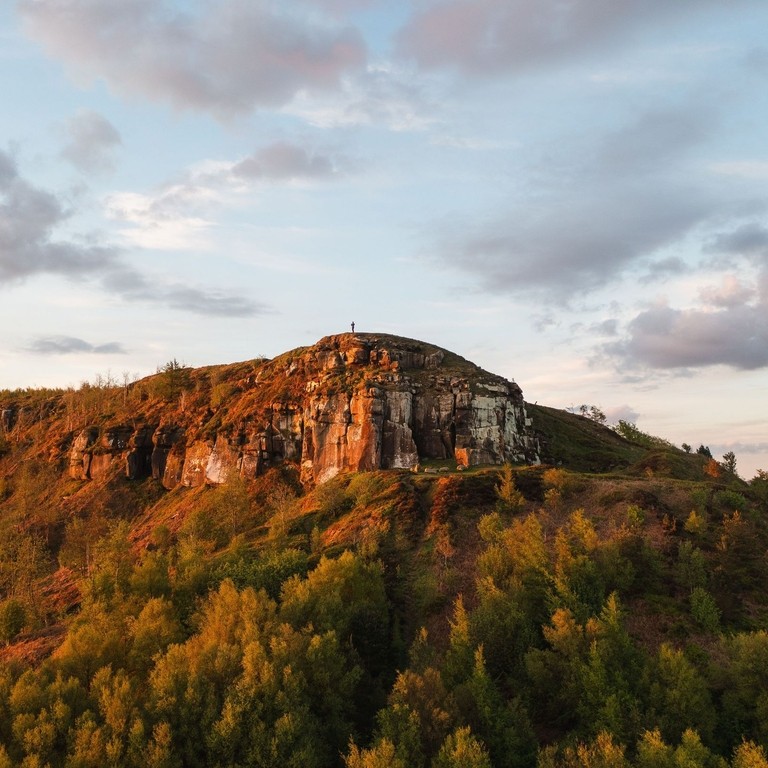 A lone person stands on a cliff top with forest below