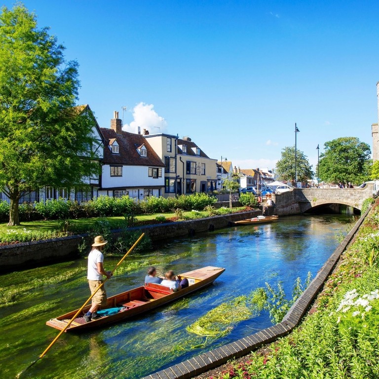 A man and a couple in a punt on a small river surrounded by gardens and houses