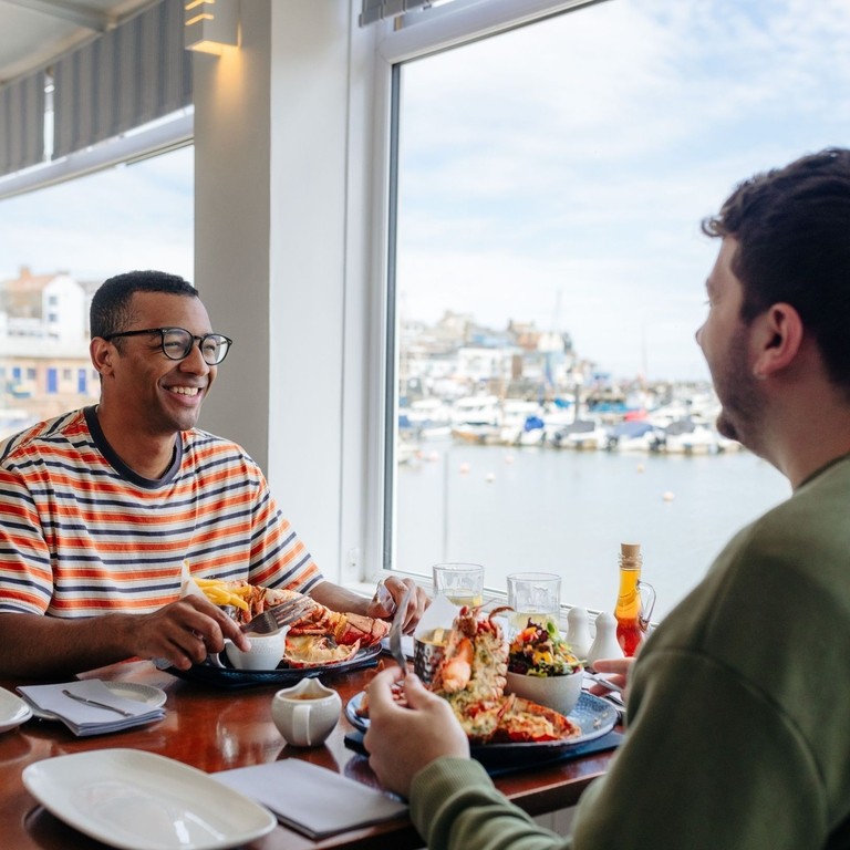 Two men eat lobster in a restaurant with a harbour view