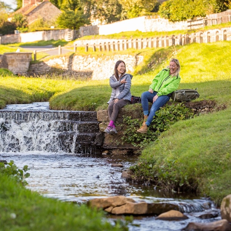 Two women sat on the banks of a stream, in a village