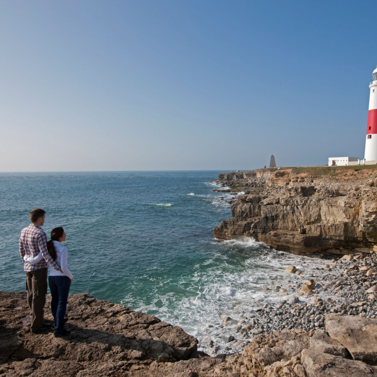 Couple standing on a cliff by the sea looking at a red and white lighthouse 