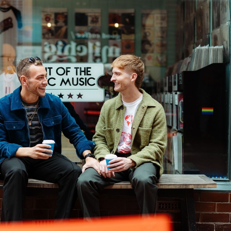 Two men sit with take away drinks in front of a shop