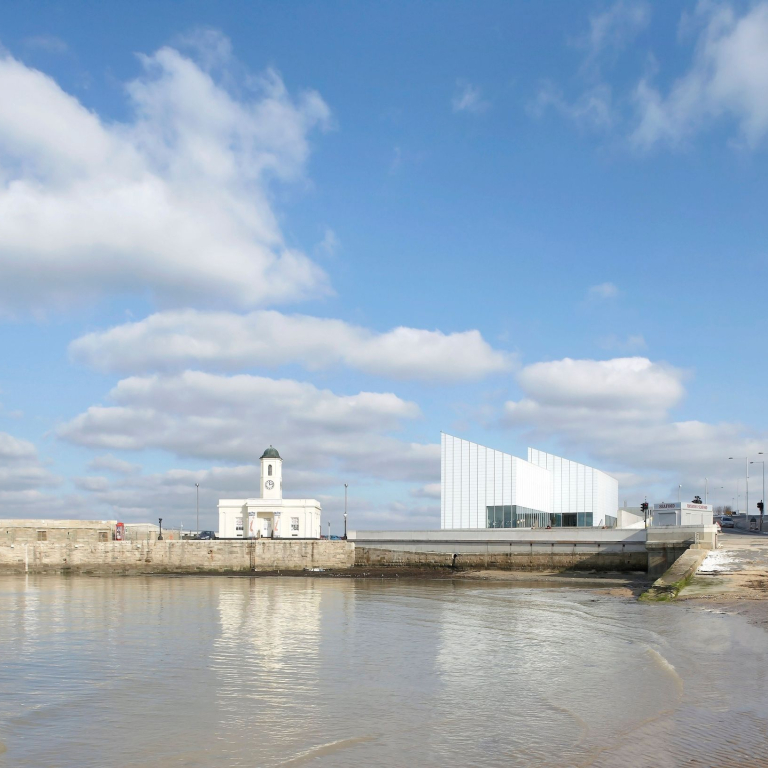 Modern building overlooking a harbour and beach, a row of townhouses in the background