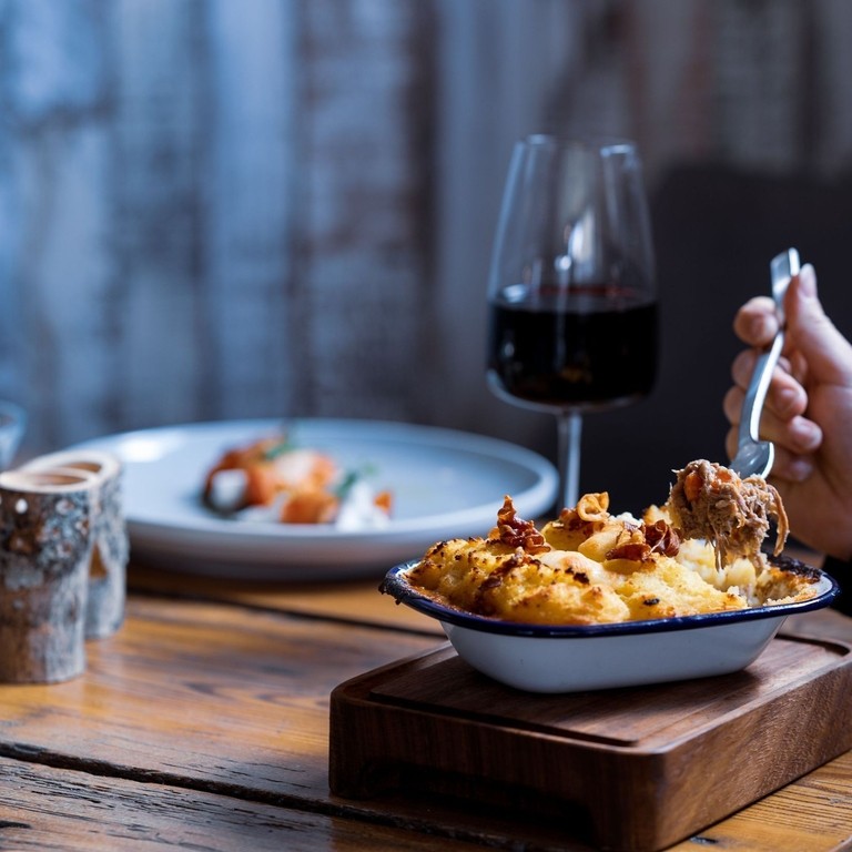 View of arm eating a mashed potatoes topped dish with two glasses of red wine and another dish of food in soft focus in the background