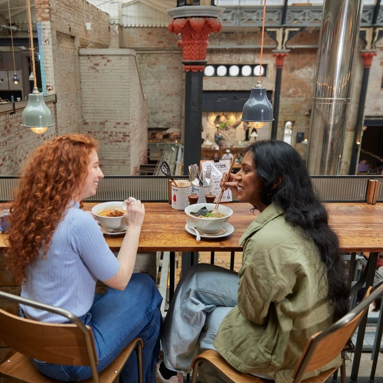 Two women enjoying lunch inside a large food hall.